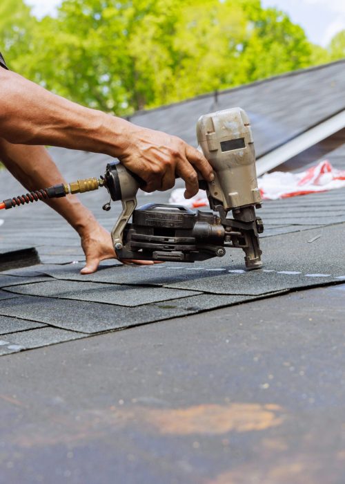 In addition to using an air pneumatic nail gun, roofer installs new asphalt bitumen shingles