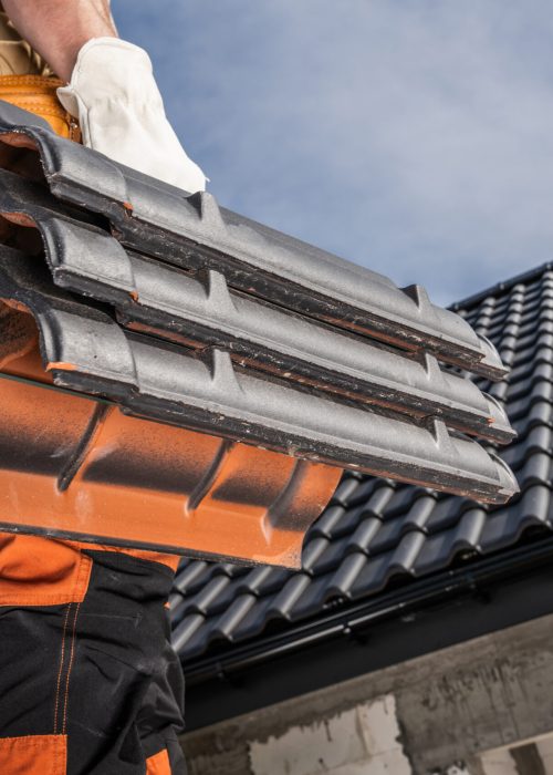 Caucasian construction roofer worker is standing on a roof, holding ceramic roof tiles.